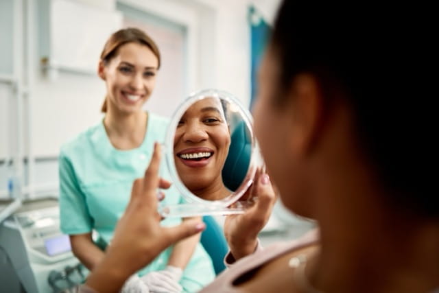 A dental patient examines her smile in a mirror while a provider looks on