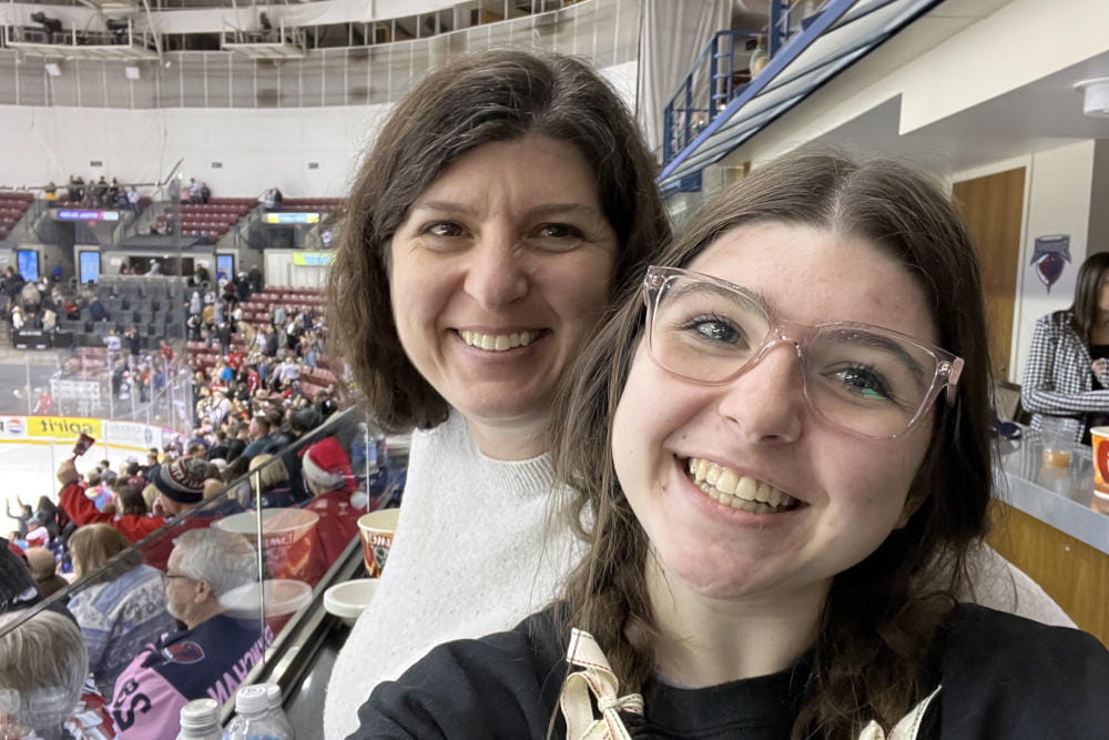 An 18-year-old woman and her mother smile at the camera from the stands of a packed hockey stadium