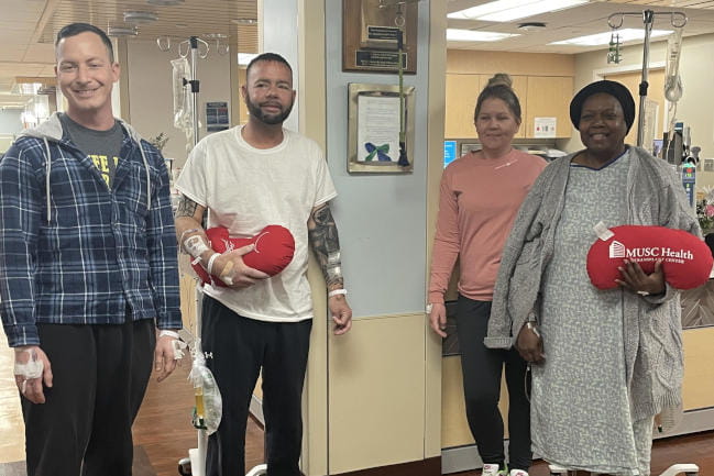 Four people stand in a hospital waiting room, some with IVs attached. They are holding plush pillows shaped like kidneys with the MUSC Health logo printed on them.
