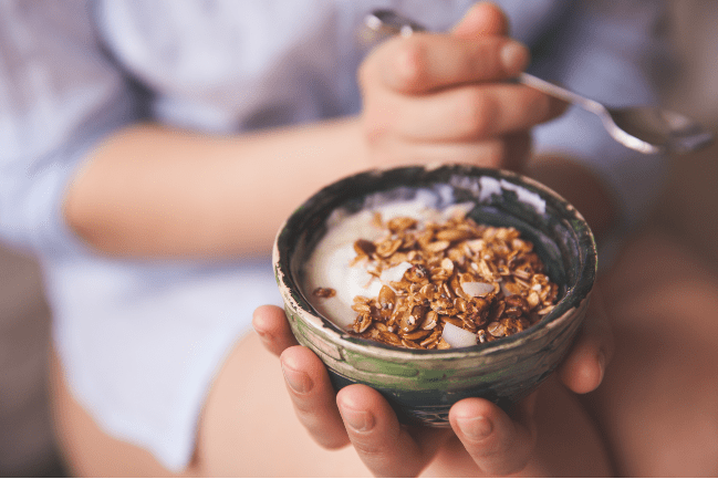Close up of a person's hands holding a small bowl of yogurt topped with granola and a spoon.