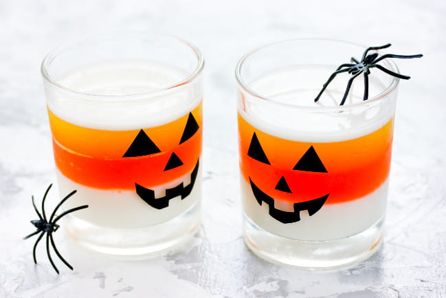 Two drink cups designed with jack-o-lantern faces. Two fake black spiders are propped against them.