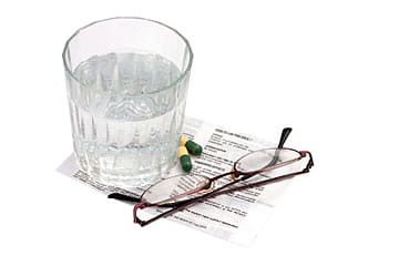 Photo of a glass of water, two capsule pills, and a pair of glasses on top of a sheet of medication instructions.