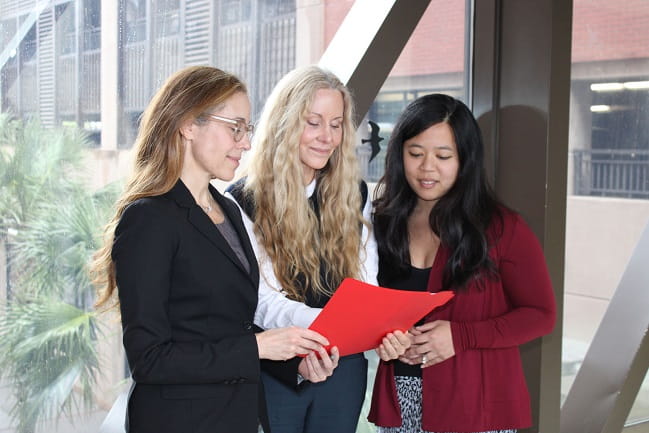 Three women looking at a binder in front of a large window. 