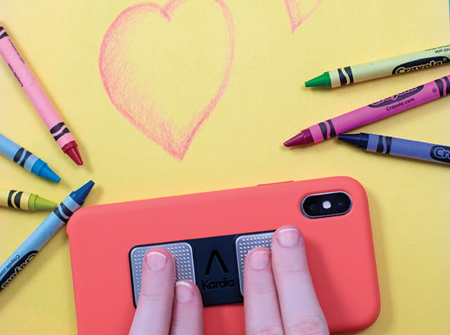 Image of a child's fingers on an electronic device surrounded by crayons scattered on a yellow background with a heart drawn on it.