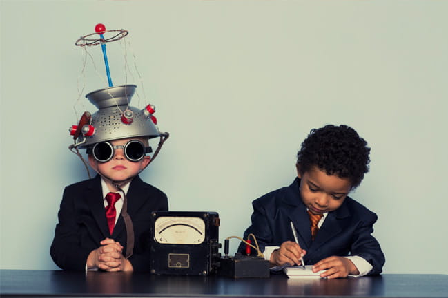 One child sits with a colander on his head and a radio next to him while another child pretends to take notes.  