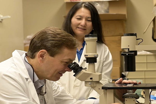 Charlie Strange, M.D., looking into a microscope while Hongjun Wang, Ph.D., looks on