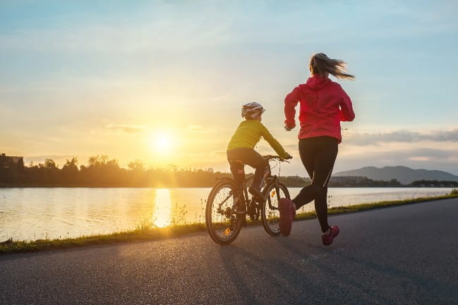 Adult jogging next to a child that is riding a bicycle.