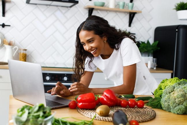 A smiling woman in a kitchen looking at a laptop with vegetables next to her.