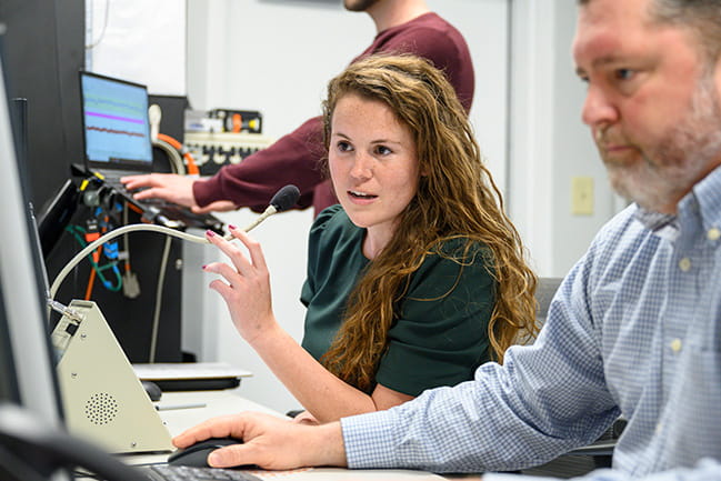 A woman speaks into a microphone while collaborating with colleagues in a research room.