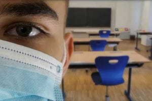 Half of a face wearing a mask with desks in the background in a classroom setting.