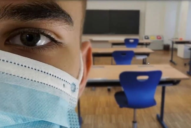 Half of a face wearing a mask with desks in the background in a classroom setting.