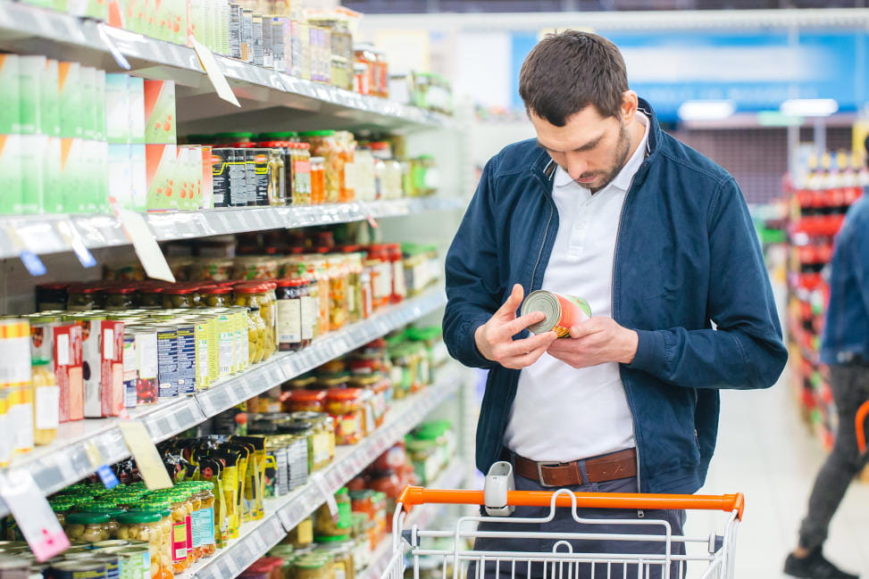 	At the Supermarket: Handsome Man Uses Smartphone and Looks at Nutritional Value of the Canned Goods. He's Standing with Shopping Cart in Canned Goods Section.