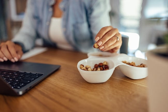 A woman uses a computer while snacking on a bowl of nuts.