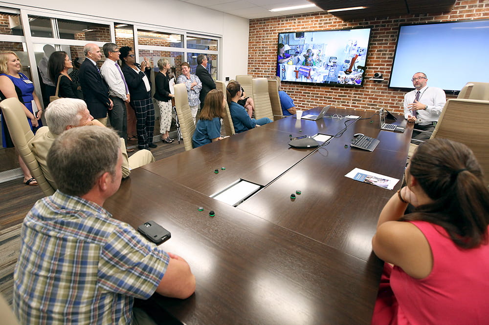 people gather around a conference room table to watch a doctor interact with a remote patient through a large video screen