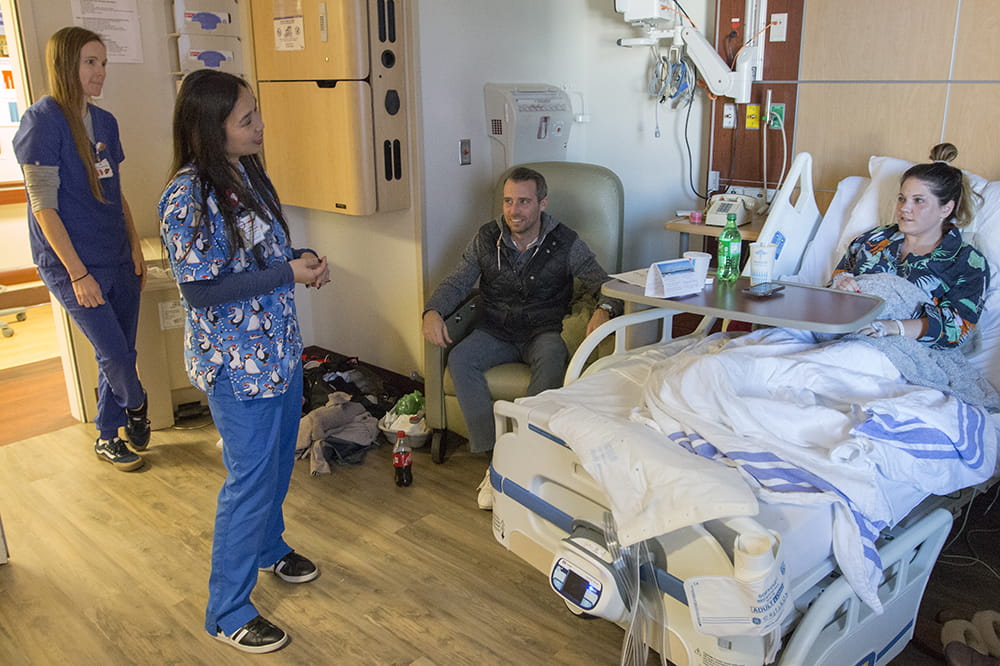 two nurses stand and talk to Laura Hoover, who is in her hospital bed. Her husband looks on from a chair nearby
