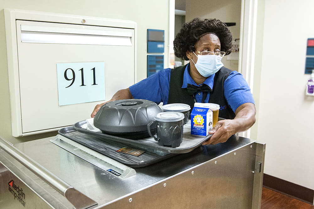 A woman takes a tray of food from a stainless steel cart to bring to a patient's room