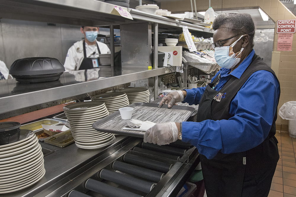 A woman in a commercial kitchen prepares a tray as the cook on the line behind her looks on