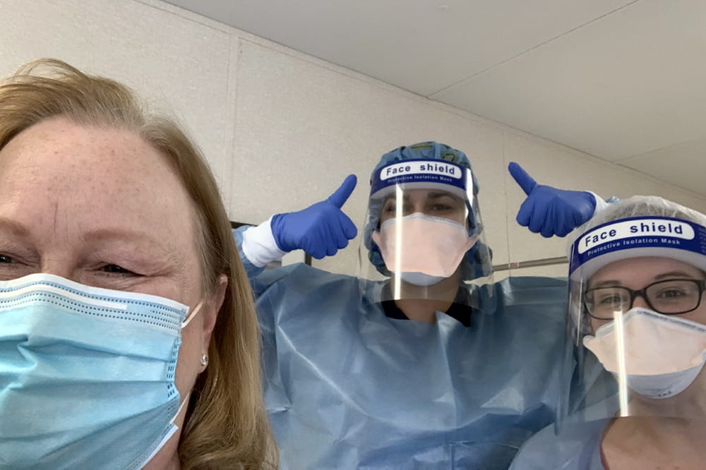Three women post for a selfie. The patient wearing a mask and the nurses wearing gowns, gloves, masks and face shields. One nurse gives two thumbs up. 