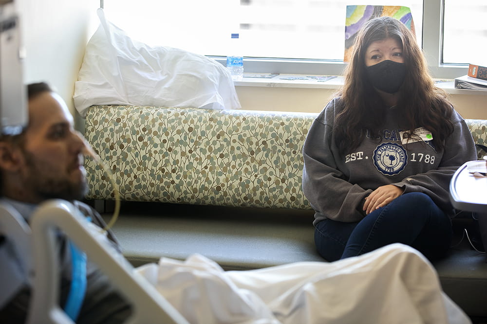 A woman sitting on a couch in a hospital room looks over at her husband