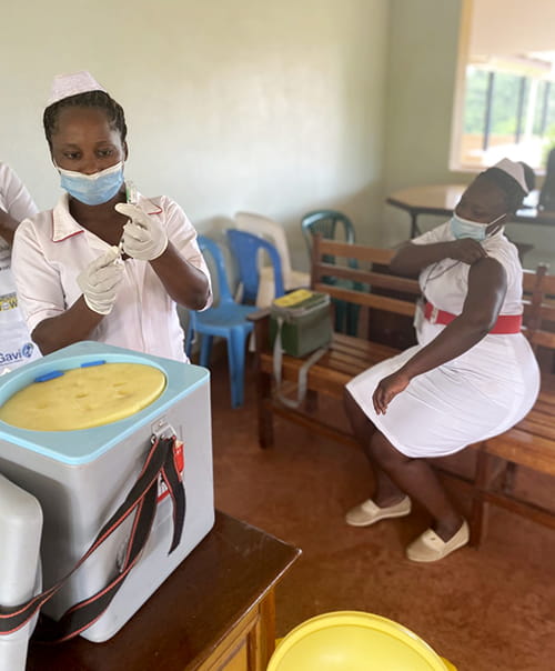 two nurses in traditional white uniforms in a clinic, one sitting and rolling up her sleeve, the other with a needle