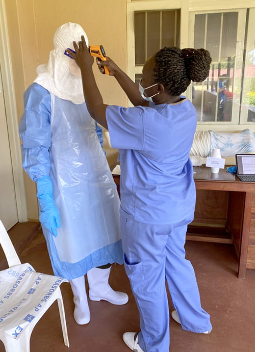 a health care worker in scrubs holds a forehead thermometer up to another worker completely encased in personal protective equipment