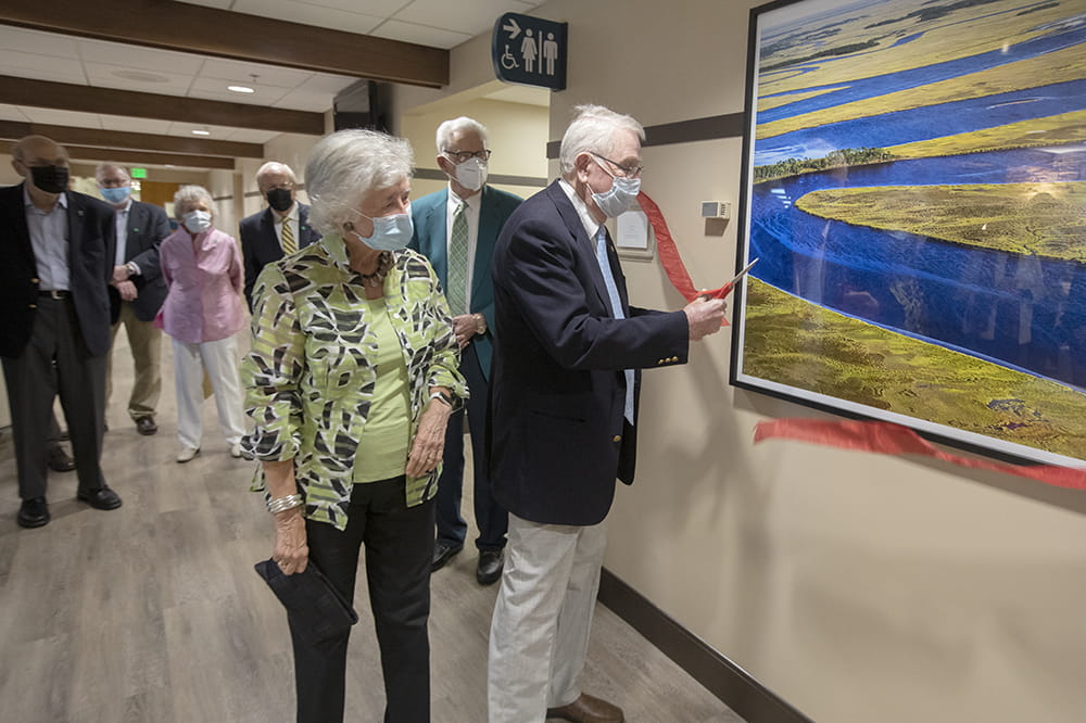 a man cuts a red ribbon taped to a wall in front of a large photograph of a Lowcountry creek