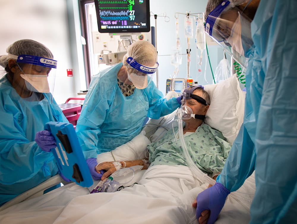 A man who would eventually die from COVID is surrounded by nurses, one of which is holding his hand