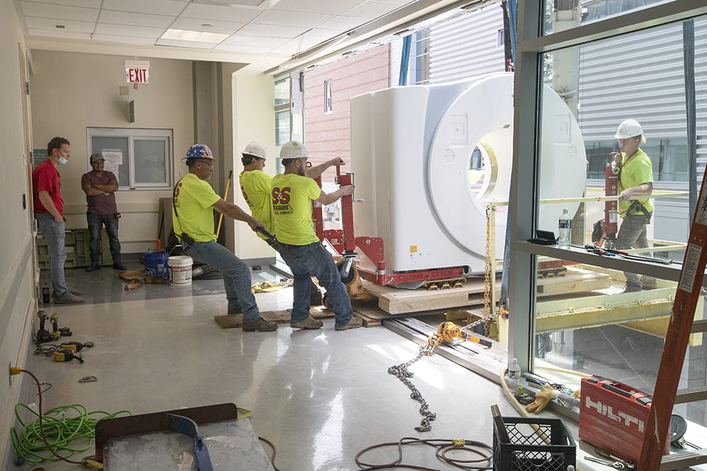 workers guide a CT machine on an oversized dolly through an open window