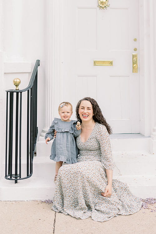 a young woman sits on a doorstep with a toddler girl standing next to her