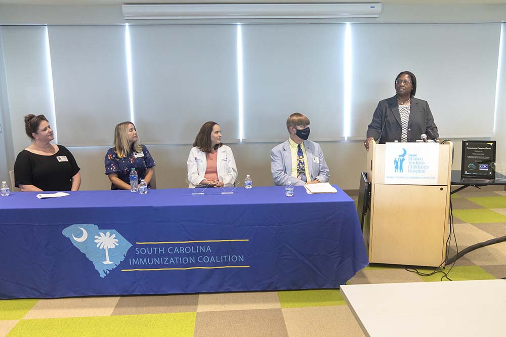 Three women and one man sit at a table covered by blue cloth, listening to Youlanda Gibbs, who is standing at a podium.