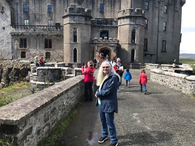 a woman in jeans and jacket stands at the long stone entrance to a castle