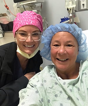 a woman in hospital gown and blue hair cover in hospital bed takes a selfie with hospital worker