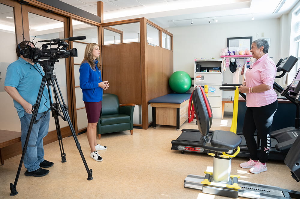 a TV cameraman and reporter talk to a woman in a pink shirt in a gym-like space