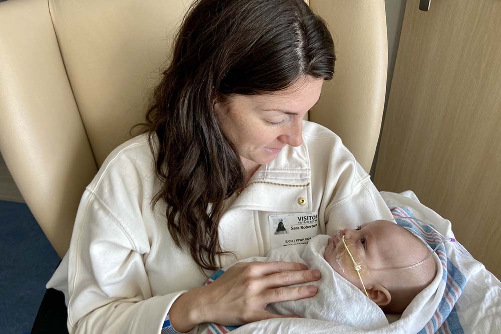 Sara and Liam Robertson. Sara sits in a chair wearing a white top with a visitor sticker on it holding baby Liam, who has a tube in his nose.