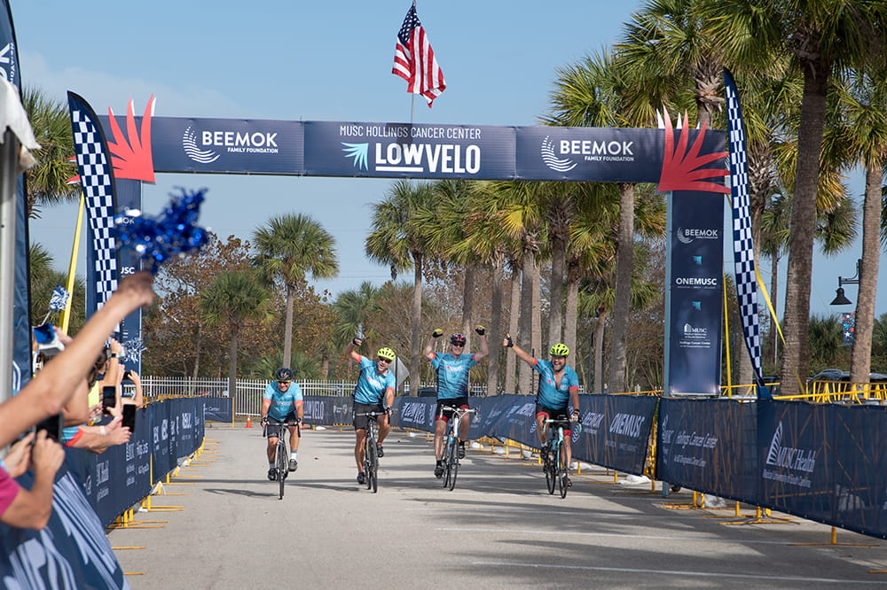 four bicyclists cross under the finish line with the American flag against a blue sky