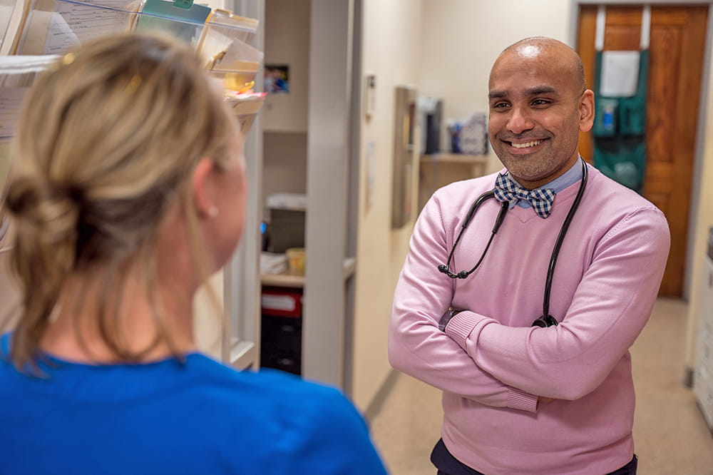 a doctor in bright pink sweater and blue bow tie speaks with a nurse in a hallway