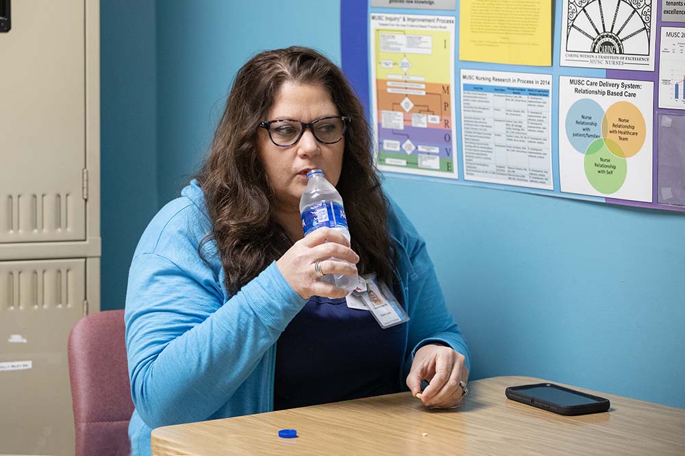 Woman with long dark hair wearing a blue jacket drinks from a water bill as she holds a vitamin.