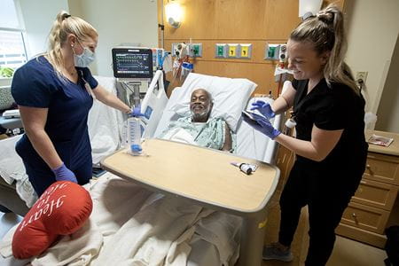 A man smiles from his hospital bed as two nurses tend to his needs