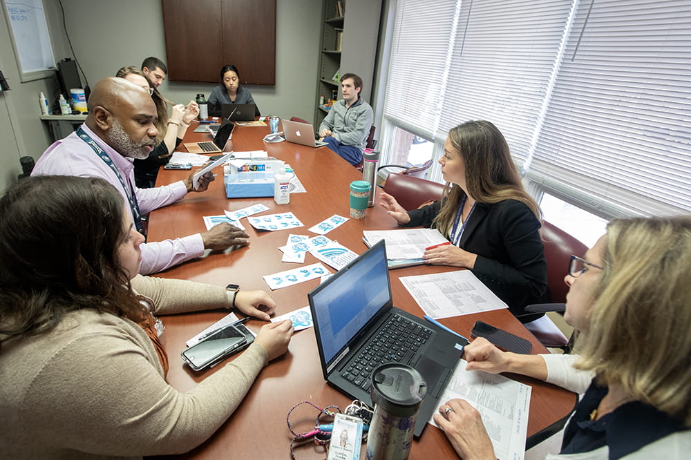 Eight people seated at a table with laptop computers and paperwork in front of them. Some have coffee cups.