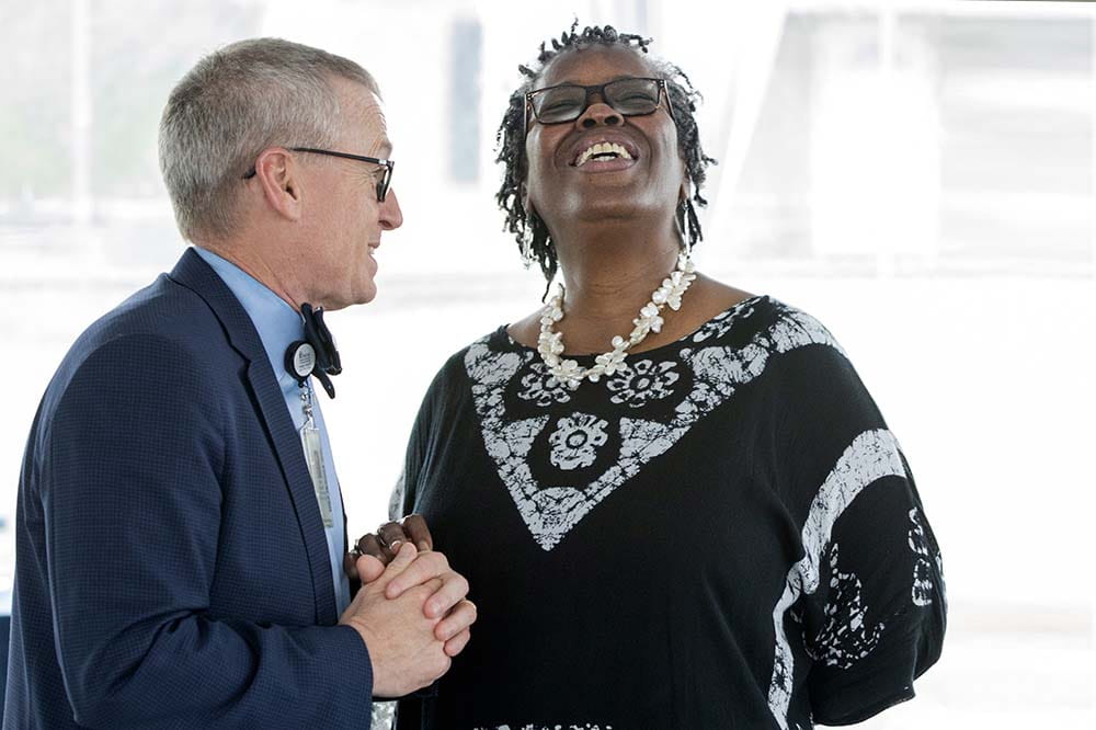 A smiling man in a suit, bowtie and glasses laughs with a woman who is also wearing glasses and a black and white blouse. They look like they are celebrating.
