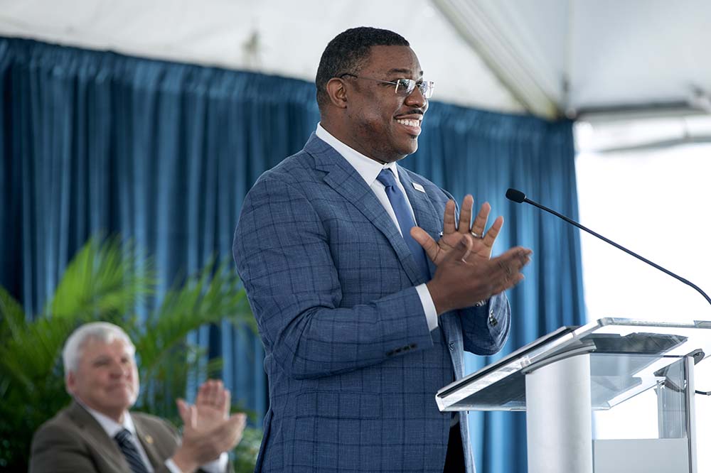 Surgeon in a blue suit and glasses smiles while he's speaking at a podium.