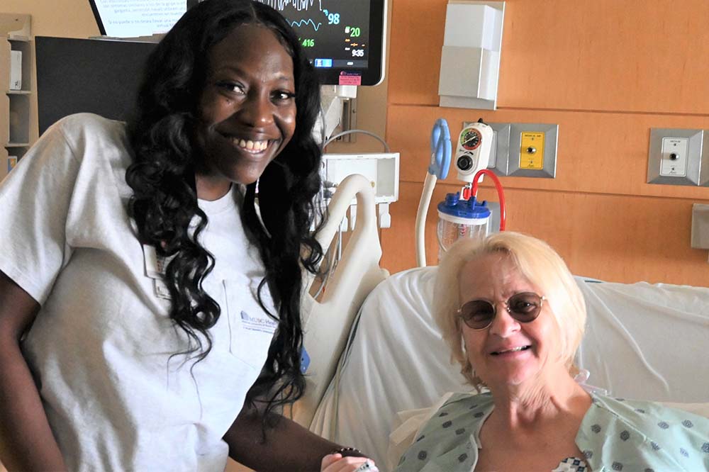 A patient in a hospital bed wearing glasses smiles while posing with a patient care tech who has long black wavy hair and is wearing a white t-shirt.