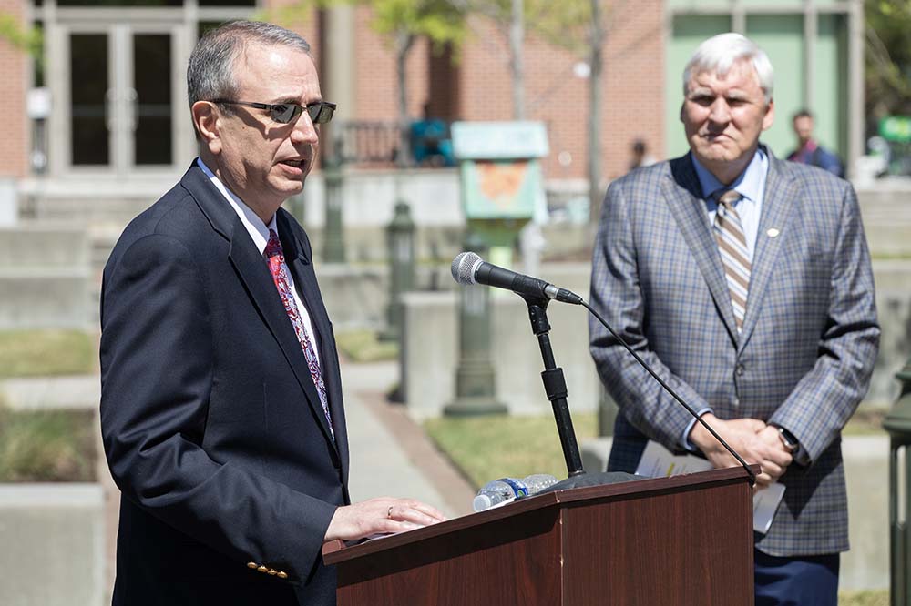 A man in a suit stands at a podium while another man in a suit listens. They are outside.