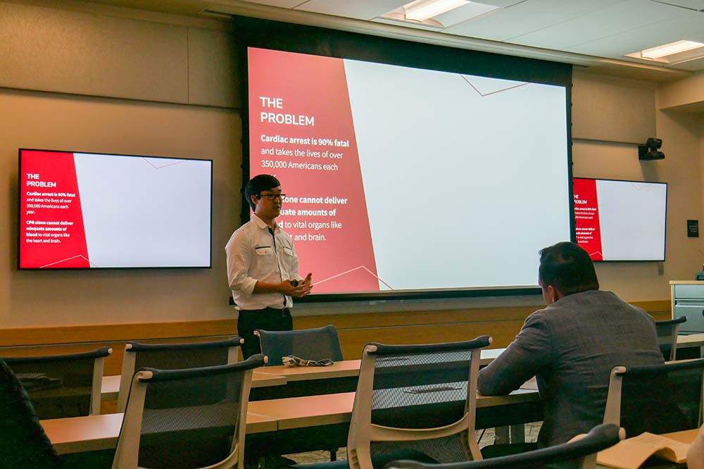 Man stands by a screen with the words "The problem. Cardiac arrest is 90 percent fatal and takes the lives of over 350,000 Americans each year." Another man, seated, is listening.