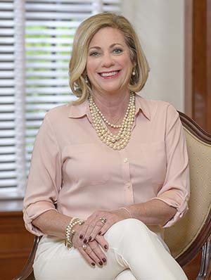 Woman with shoulder length blonde hair wearing a pink shirt and white pants smiles while seated.