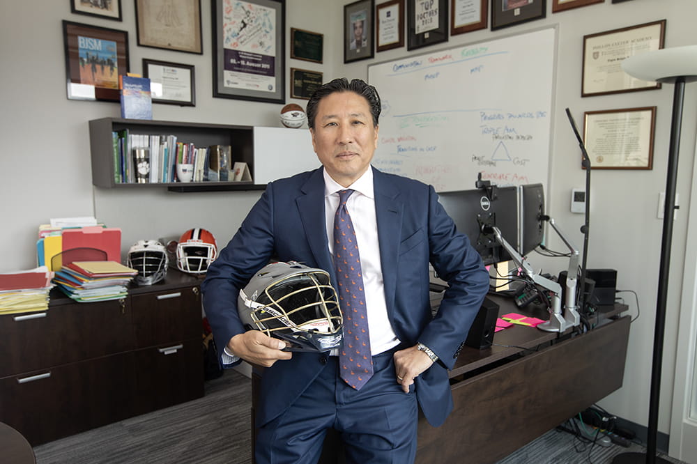 Man in a suit holds a lacrosse helmet as he leans against a desk. There are plaques behind him.