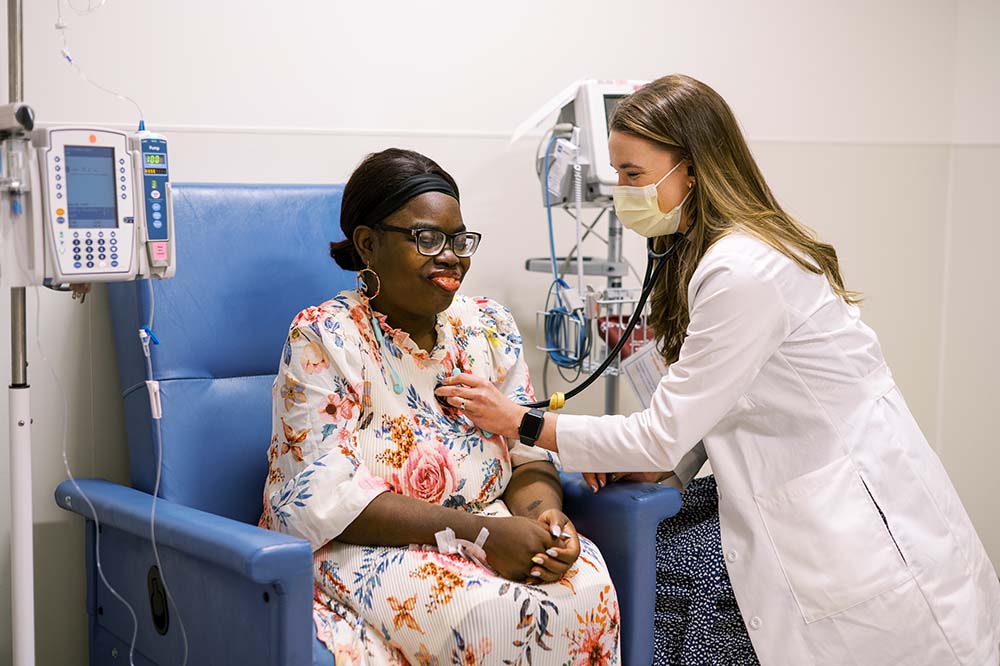 A woman wearing a floral dress and glasses is seated while another woman wearing a white lab coat performs a medical check.