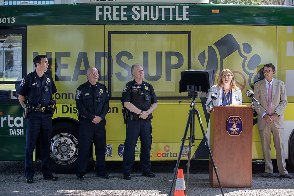 Three police officers and a city councilman flank a female surgeon speaking at a dais in front of a bus with the Heads Up signage on its side