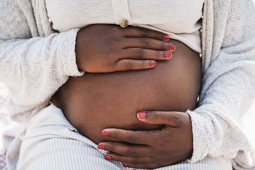 Woman holds her pregnant belly. It is bare. She is wearing a white/cream colored outfit.
