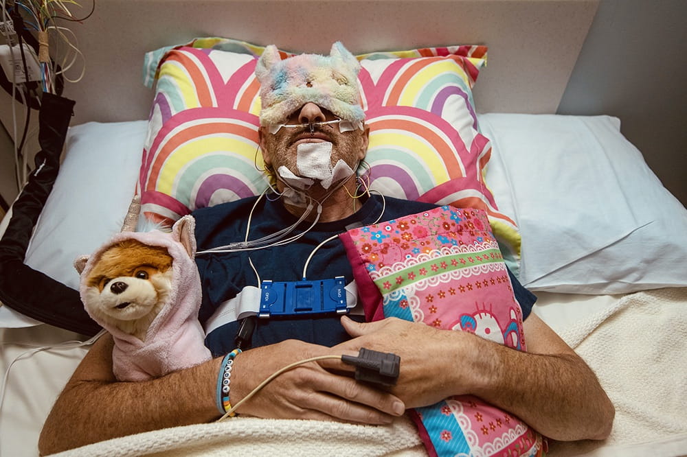 A man in bed wearing a sleep mask, holding a stuffed Pomeranian wearing a unicorn onesie and a hot pink Hello Kitty pillow